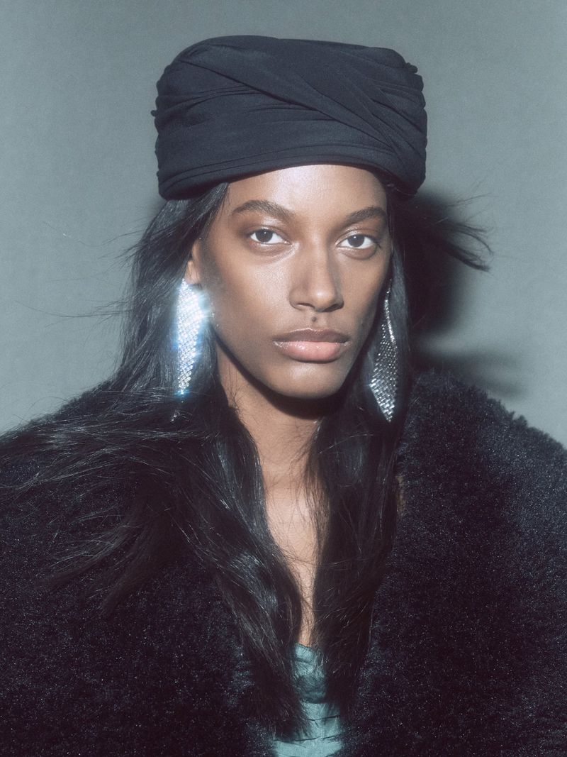 Striking studio portrait of a Black woman with a black turban and shimmering earrings, wearing a dark fur coat against a grey backdrop.