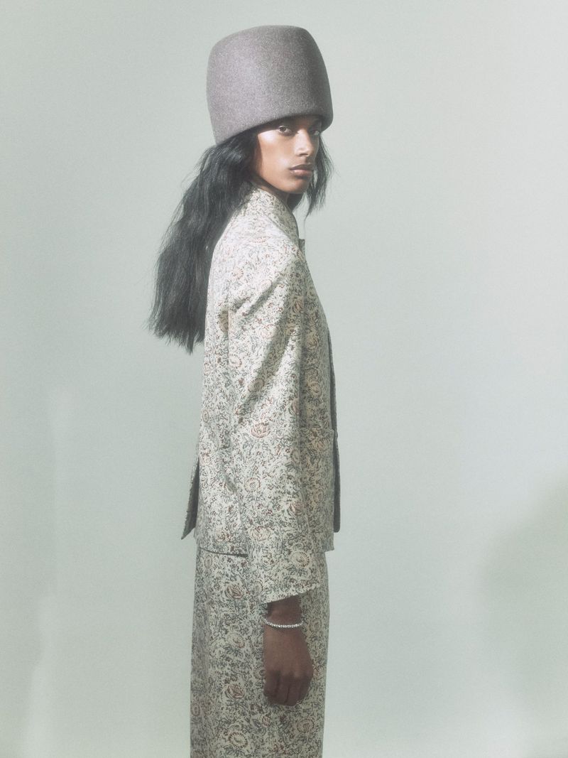 Profile shot of an adult female wearing a patterned beige jacket and skirt with a grey fur hat against a light backdrop. 