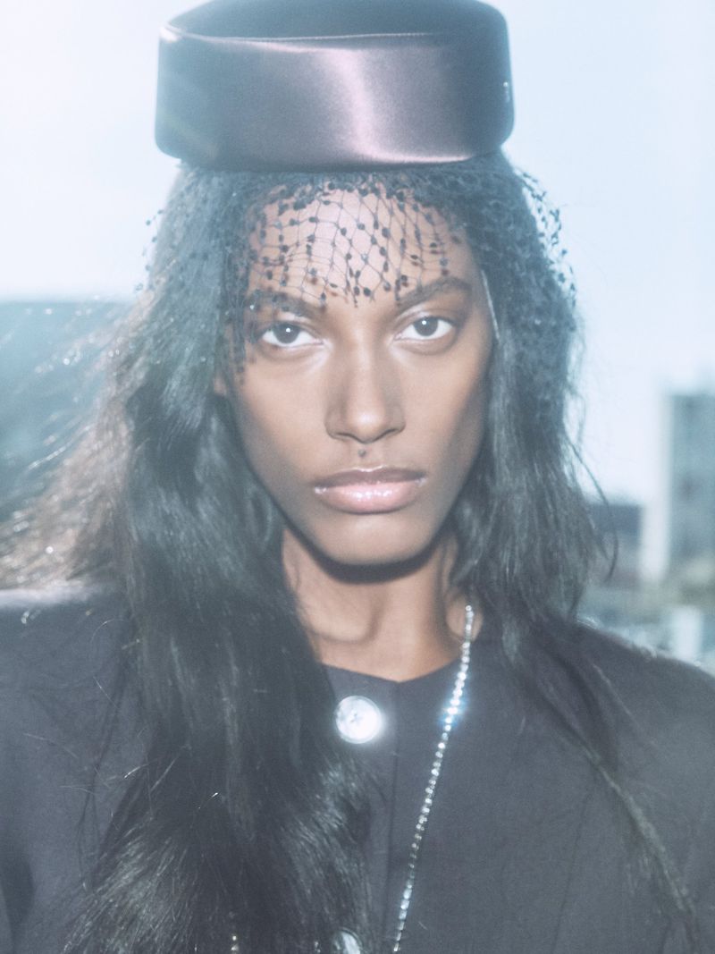 Elegant portrait of an African-descent woman wearing a netted headpiece and dark blazer in an urban outdoor setting.