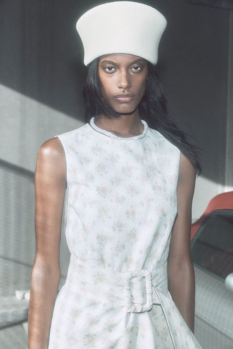 Fashion portrait of an adult woman wearing a white floral-patterned sleeveless dress with a white hat inside an indoor setting.