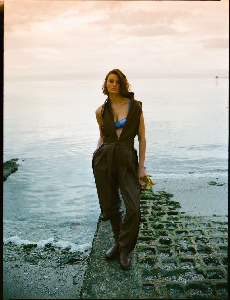 A woman in a brown jumpsuit and boots stands on a concrete structure with water and a sky in the background, creating a fashion editorial.