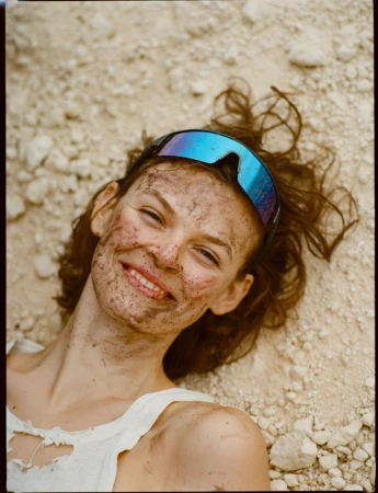 A smiling woman covered in dirt is reclined on a rocky surface, wearing goggles; captured with a medium shot in outdoor lighting.