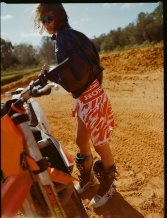 Young adult poses with a dirt bike in an outdoor setting, wearing a jacket, skirt, and boots; natural light, medium shot.
