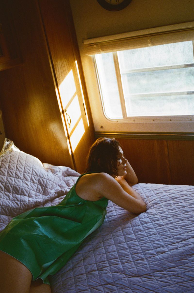 Adult female rests on a patterned bed indoors, wearing a green satin dress, near a window with sunlight, creating a warm, serene scene.