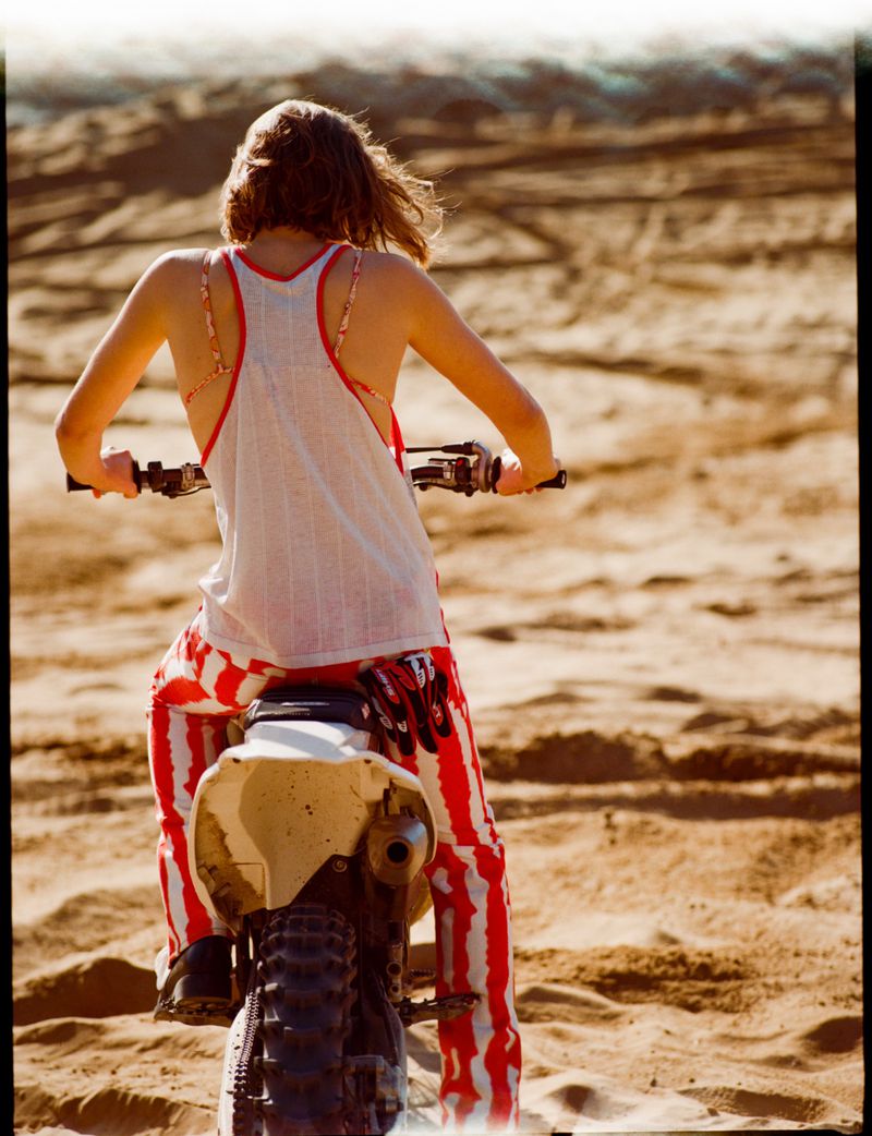Back view of a woman riding a dirt bike across a tan sand dune. She wears a white top and red and white striped pants.