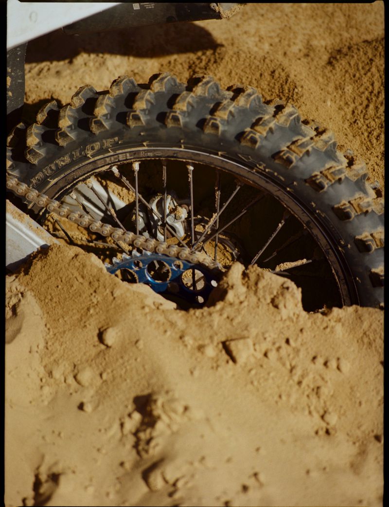 A motocross bike wheel is partially buried in tan sand, showing the tire tread, spokes, chain, and sprocket.