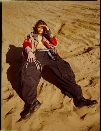 A woman in a jacket and oversized pants sits in a desert with a hand over her eyes against a sandy, dune-filled background.