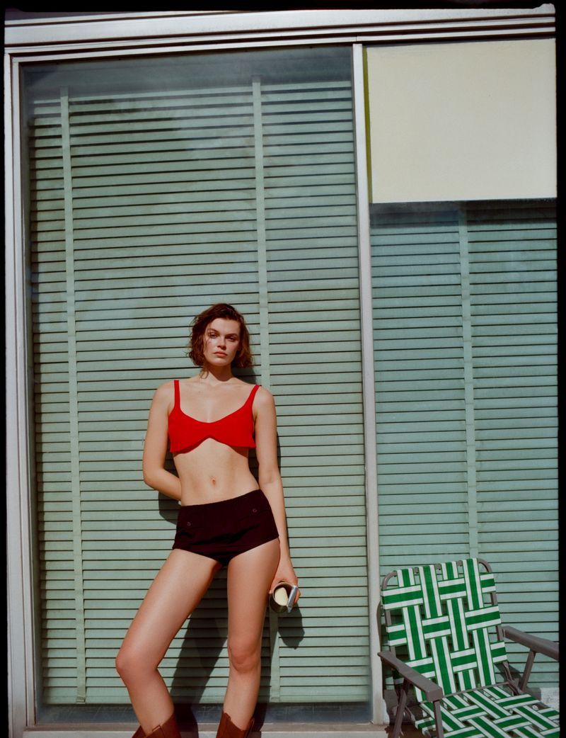 A woman in a red bikini top and brown shorts poses in front of a window with horizontal blinds, wearing brown cowboy boots and holding a can.