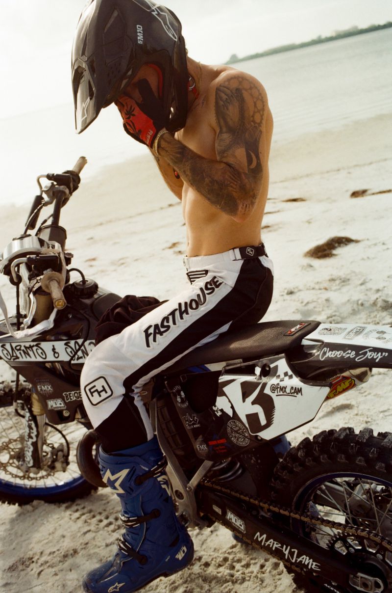 Tattooed adult male sits on a dirt bike wearing a helmet and motocross gear at the beach with a sea and sky background.