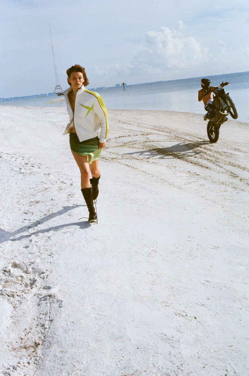 A woman in a jacket and skirt walking on a beach as a person rides a motorcycle on one wheel in the distance under a blue sky.