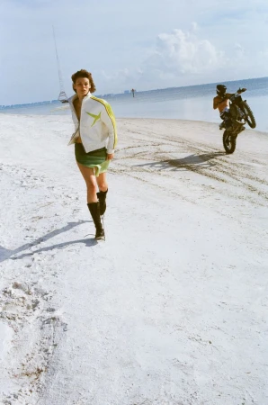 A woman in a jacket and skirt walking on a beach as a person rides a motorcycle on one wheel in the distance under a blue sky.