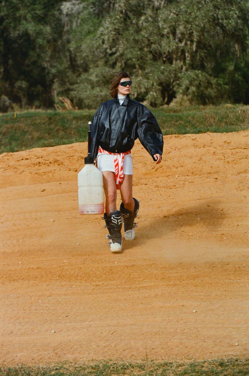 An adult female strides across an outdoor dirt field, wearing a black bomber jacket, white shorts, motocross boots, and holding a plastic jug.