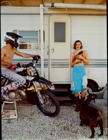 Outdoor shot of a woman holding a chicken next to a shirtless man on a motocross bike, with a goat and an RV in the background.