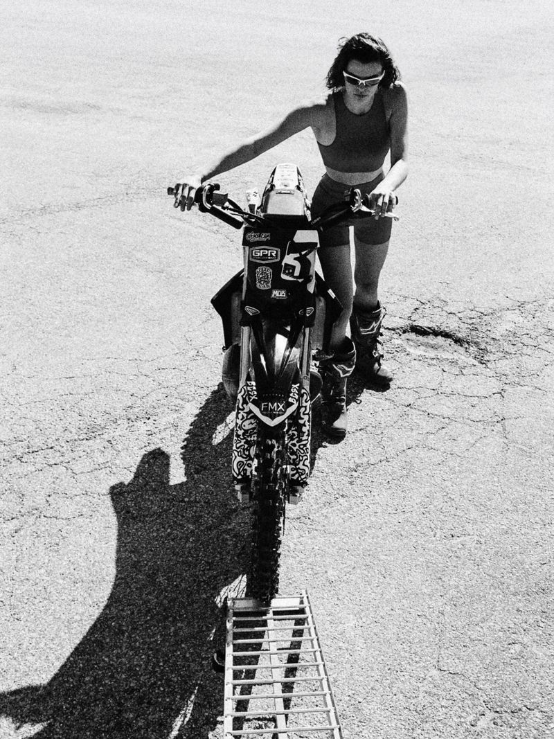 Black and white shot of an adult female with sunglasses, near a motorcycle on cracked asphalt in an outdoor setting, shot from above.