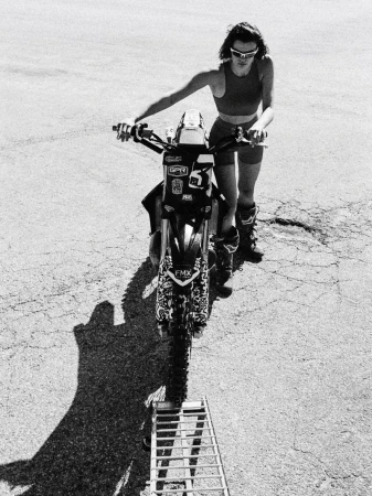Black and white shot of an adult female with sunglasses, near a motorcycle on cracked asphalt in an outdoor setting, shot from above.