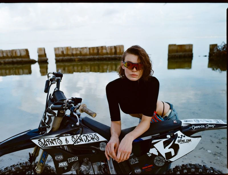 Young woman in sunglasses poses with a black dirt bike near a body of water, with a concrete structure in the distance against an overcast sky.