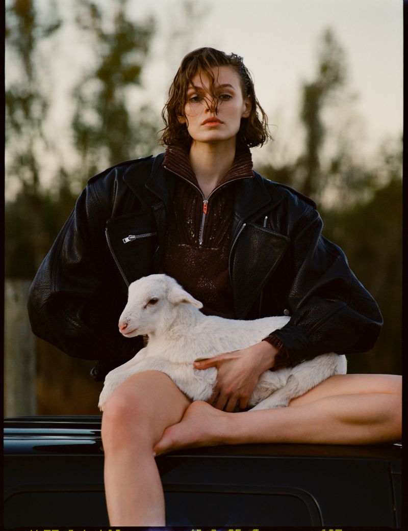 A young woman in a leather jacket poses outdoors holding a white lamb, with a blurred natural background for a fashion editorial.