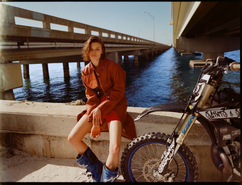 Woman in orange jacket sitting barefoot near motorcycle on concrete wall with bridge and ocean in background under blue sky.