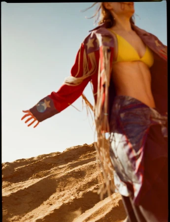 Woman in yellow bikini top and red jacket with fringe poses on sand dune against a bright blue sky on a sunny day.