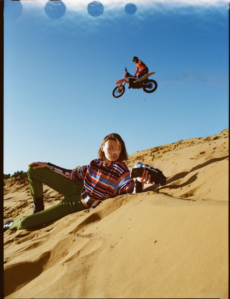 Fashion shot on a sand dune with a model, camera, blue sky, and a motocross bike jumping in the background against a blue sky.