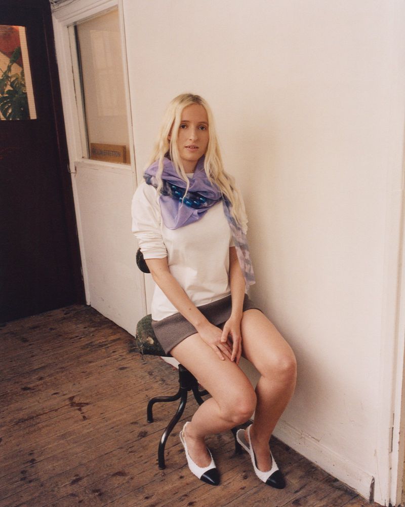 Blond woman sitting on a stool in a white shirt and taupe skirt, wearing a patterned purple scarf and two-tone heels in an indoor setting.