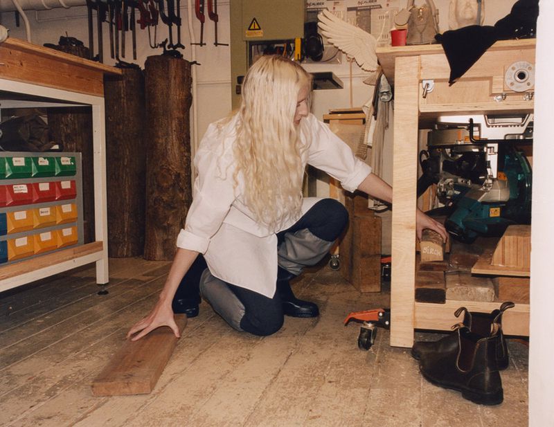Woman in white shirt and grey pants squats near workbench in wood shop with boots and wood plank on the floor of the indoor setting.