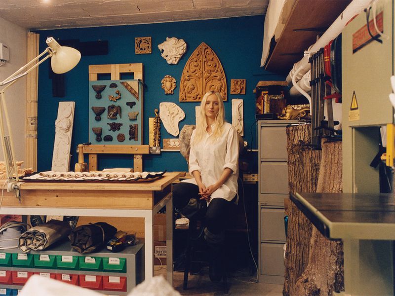 Woman in white shirt sitting in artist studio filled with wooden carvings, tools, and a teal wall with mounted artwork.