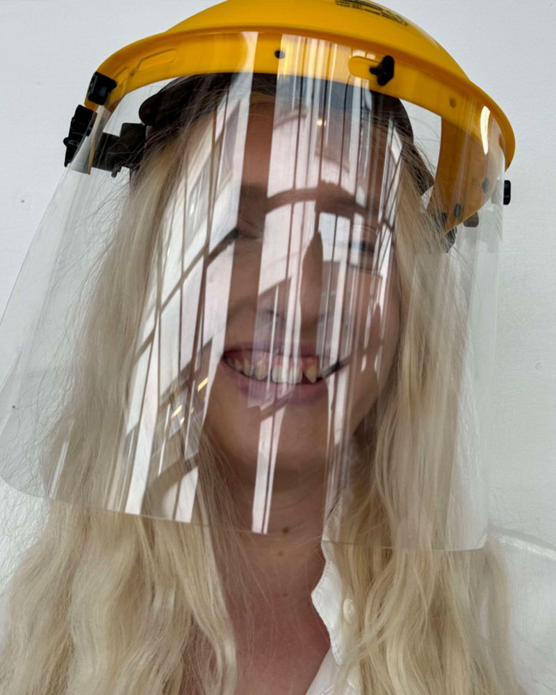 Adult female with long blonde hair smiling behind a clear protective face shield and yellow hard hat against a white background.