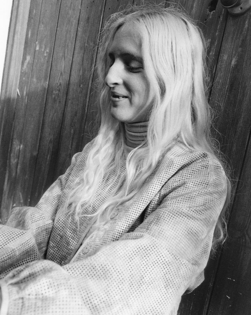 Monochrome portrait of a woman with long blonde hair wearing a patterned top against a vertical wooden paneled wall.