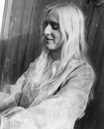 Monochrome portrait of a woman with long blonde hair wearing a patterned top against a vertical wooden paneled wall.