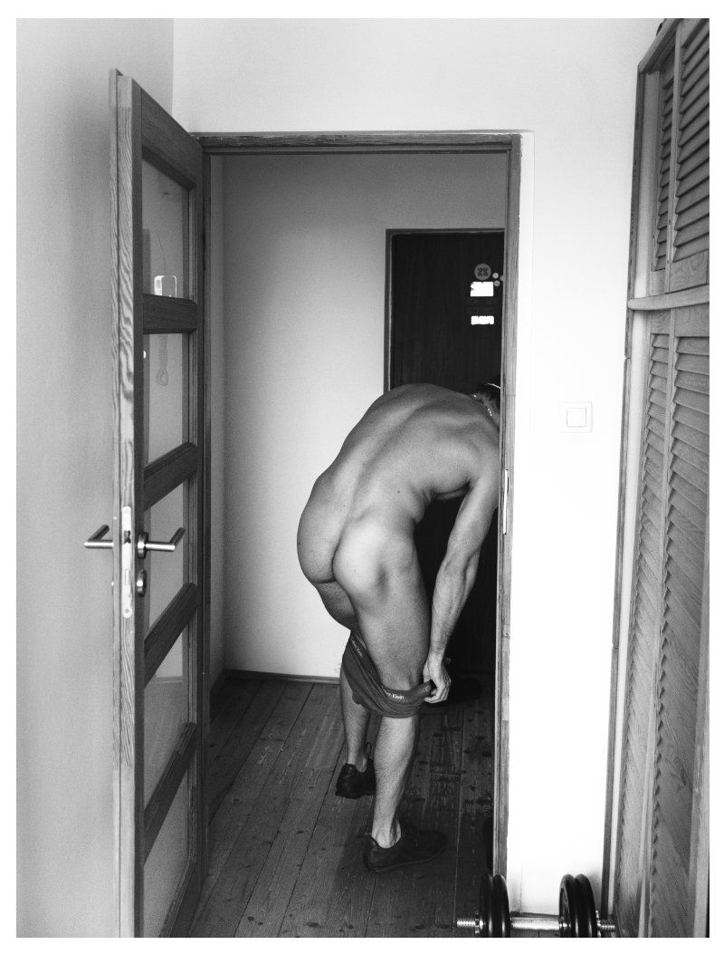 Monochrome image of a nude adult male partially visible through a doorway, pulling up underpants in an indoor setting with weights.