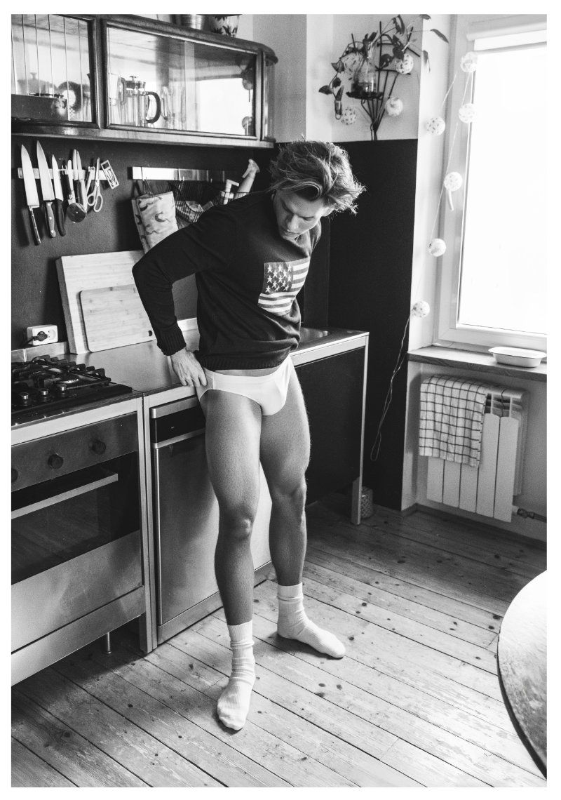 Black and white portrait of a man indoors, wearing a sweatshirt with an American flag graphic, standing in underwear and socks in a kitchen.