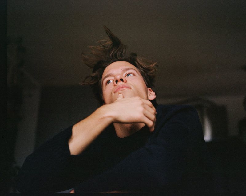 Portrait of a young adult male with brown hair, gazing upwards with a thoughtful expression in a dimly lit interior space.