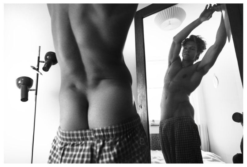 Monochrome portrait of a muscular man stretching in a mirror, wearing patterned boxer shorts, with interior and lighting details.