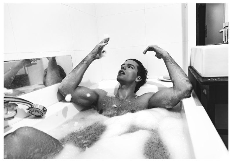 Athletic man relaxes in a bathtub filled with bubbles. He's raising his arms and looking up. The bathroom features a shower and a white tub.