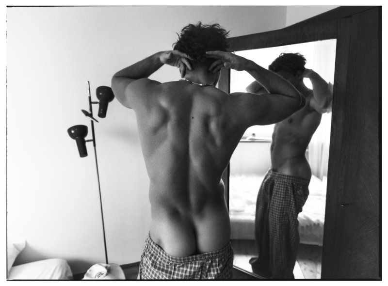 Black and white photo of a muscular shirtless man adjusting his hair while looking in a mirror in an indoor setting.