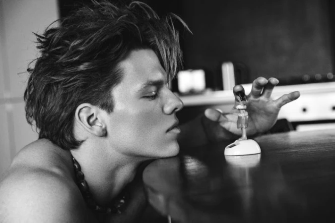 Striking black and white portrait of a young shirtless man with a beaded necklace leaning on a table, touching a small figurine.