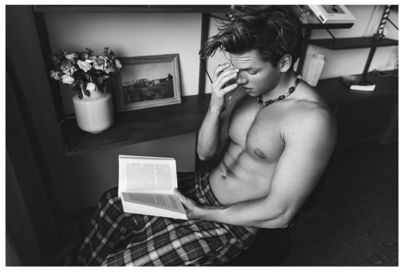 Shirtless man sits reading a book in natural light, wearing patterned pants and a beaded necklace indoors with flowers nearby.