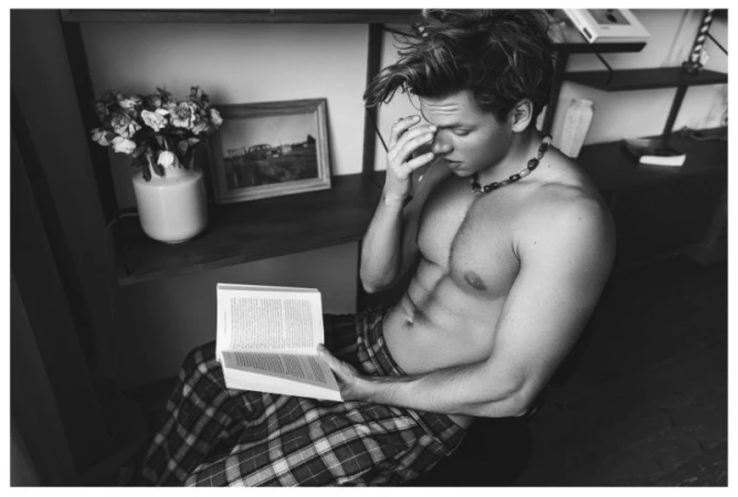 Shirtless man sits reading a book in natural light, wearing patterned pants and a beaded necklace indoors with flowers nearby.