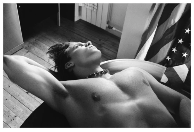 Monochrome portrait of a shirtless man lying down with arms behind his head near an American flag
