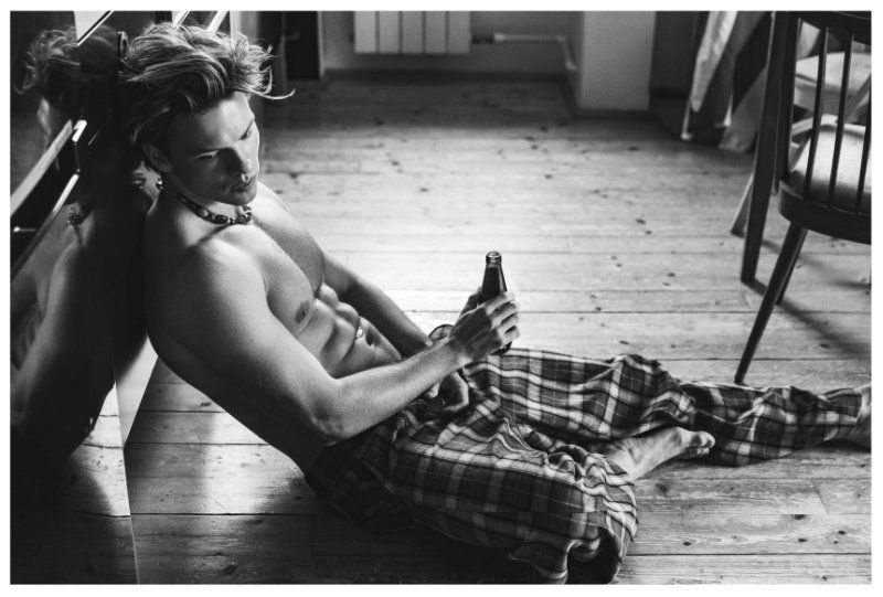 Black and white image of a man with abs, wearing pajamas, and holding a beer bottle while sitting on the floor indoors.