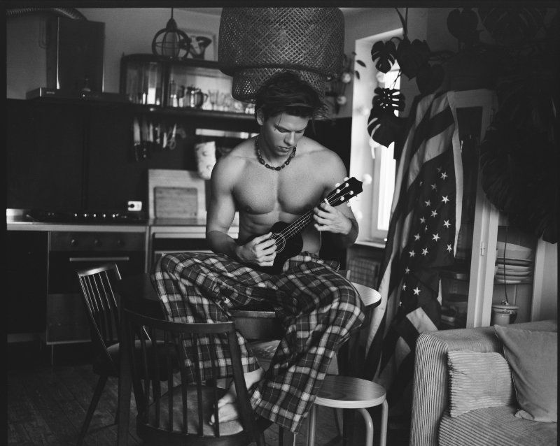 Black and white indoor shot of a fit man playing the ukulele while sitting on a stool in a kitchen, wearing patterned pants.