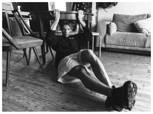 Young man sitting on a wooden floor holding onto the underside of a round table in an interior living space.