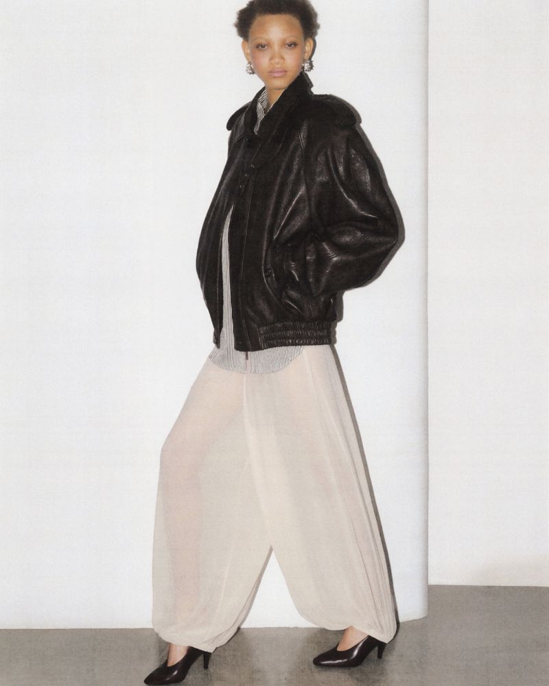Adult female fashion model poses in a studio wearing a black leather jacket and sheer, light beige harem pants with black heeled shoes.