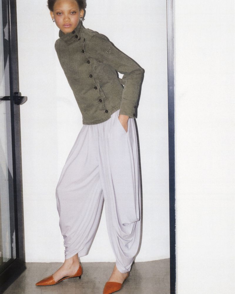 Full-length shot of a woman in high-neck jacket and harem pants with her hand in her pocket, posing in a minimalist setting.