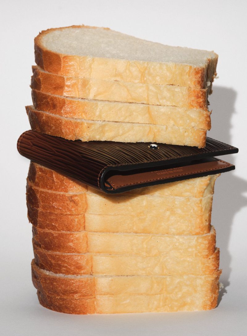 Close-up of a stack of sliced white bread, featuring a dark brown wallet placed in the middle, shot against a white background.