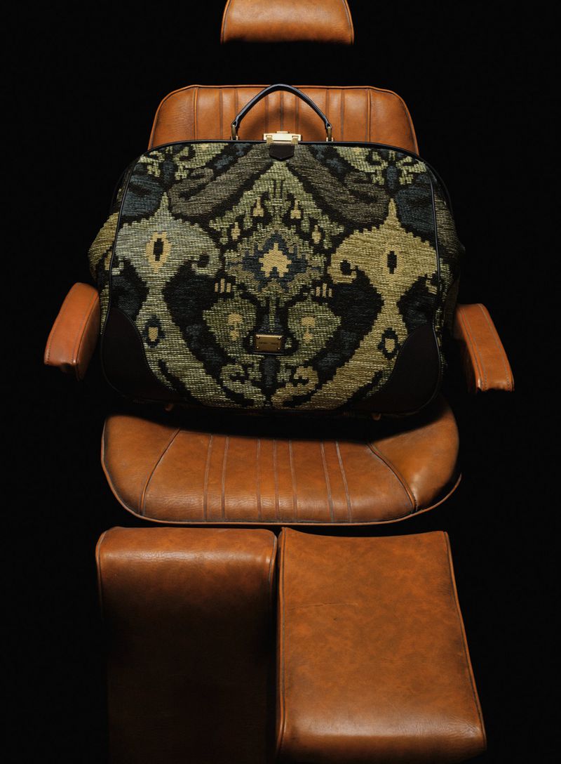 Tapestry patterned carpet bag on a leather chair against a dark background, showcasing luxury travel accessories and bespoke design.