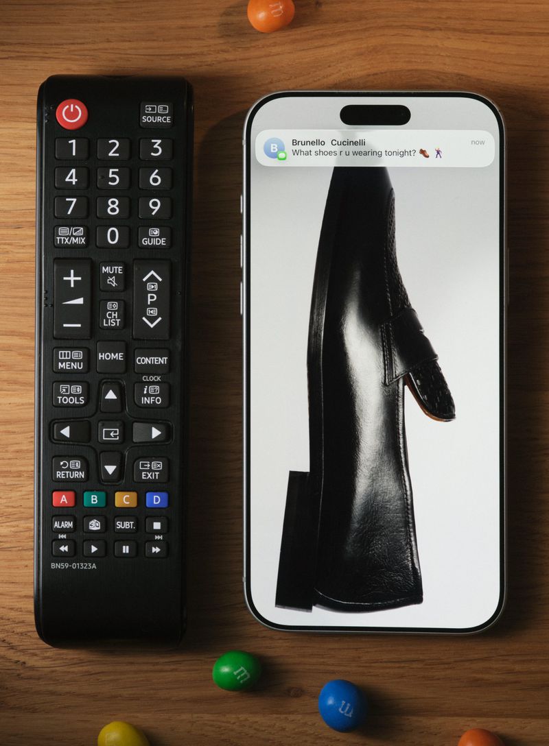 Close-up of a cell phone screen displaying a black shoe, lying next to a TV remote control and colorful candy on a wooden surface.
