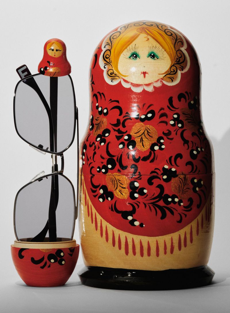Red matryoshka doll displaying eyeglasses. Small doll tops glasses, and the larger doll is in the background on a plain background.