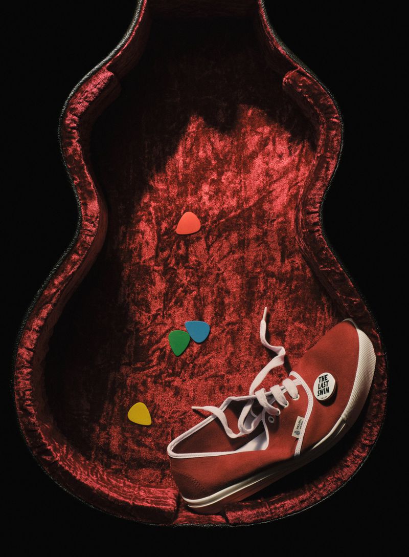 Guitar case interior with guitar picks and a red canvas shoe with white laces and sole, set against a black backdrop.